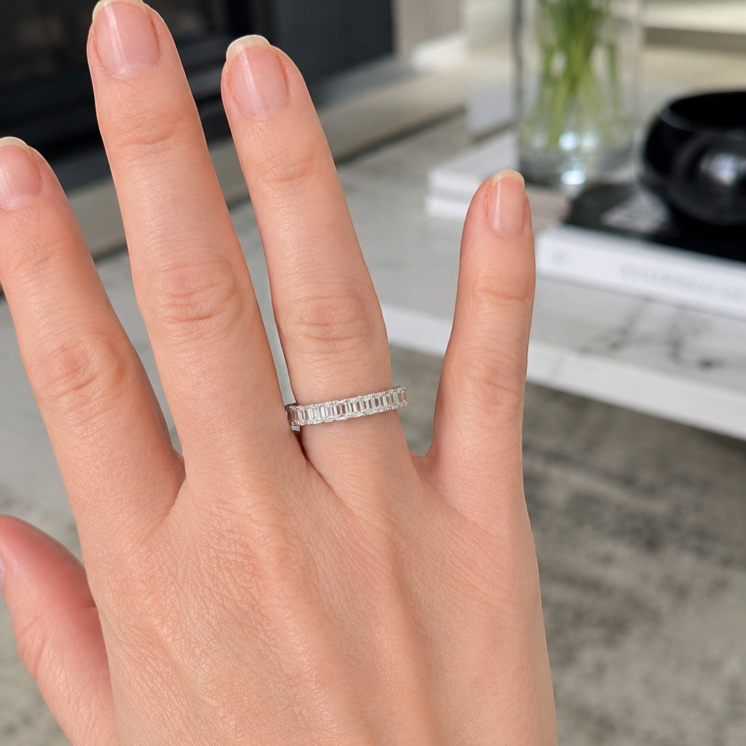 A close-up of a hand wearing a thin silver band ring with engraved text, set against a softly blurred indoor background.