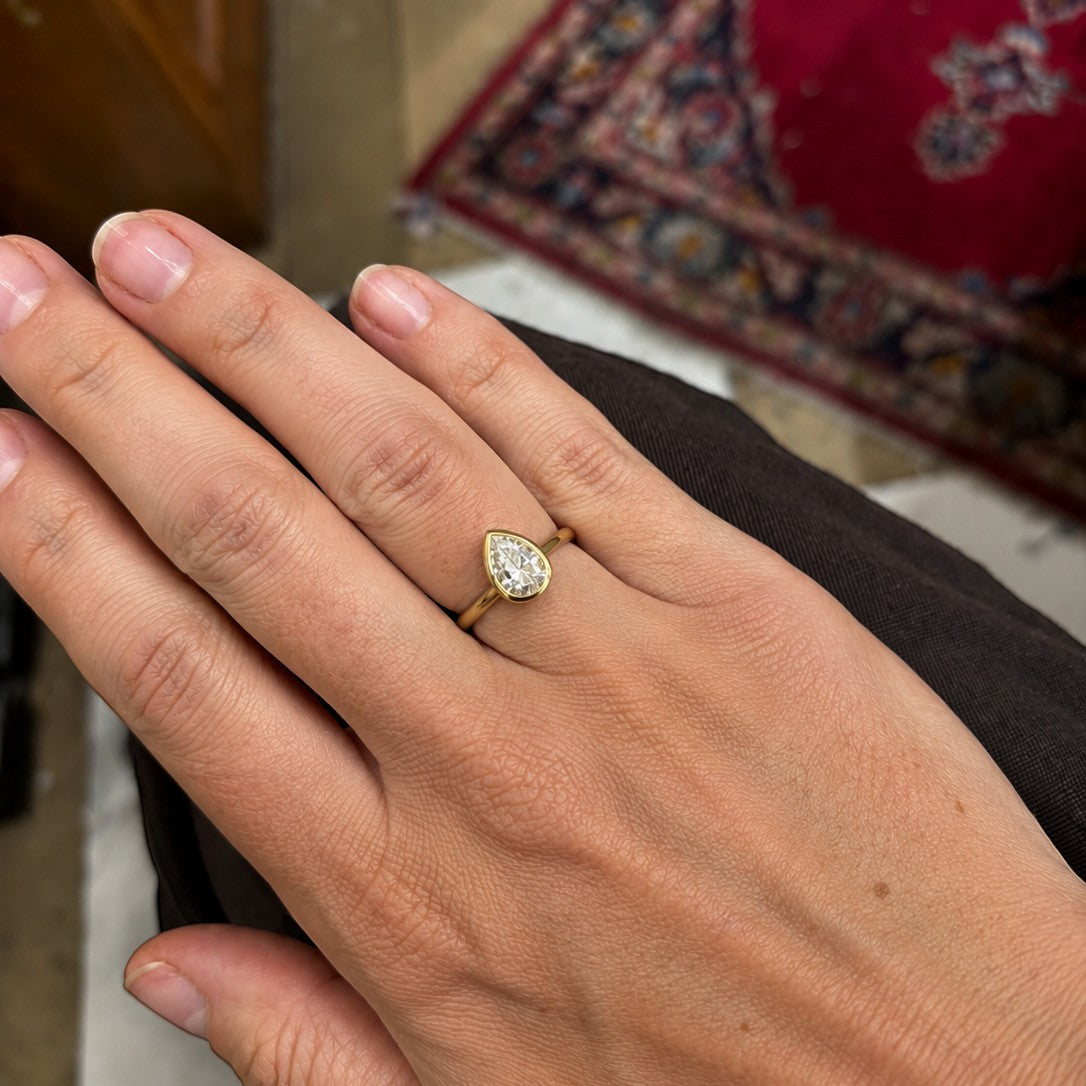 A close-up of a person's hand wearing a gold ring with a pear-cut clear gemstone, set against an indoor background with a patterned rug.