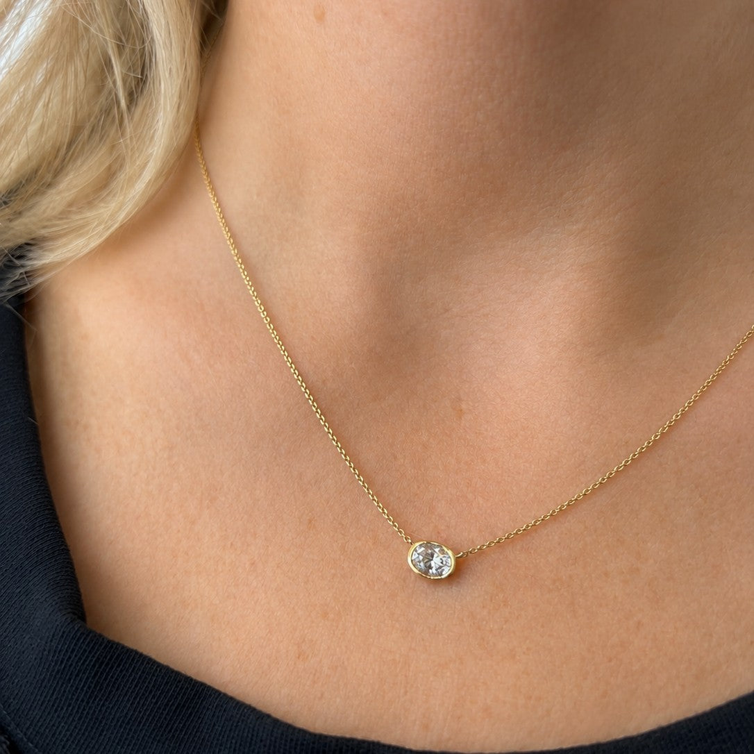 A close-up of a model's neck and upper chest wearing a delicate gold chain necklace with a small bezel-set clear gemstone pendant.