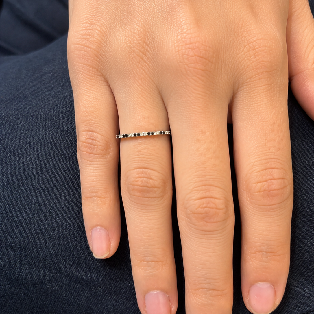 A close-up of a person's hand wearing a very thin silver band ring with small black and white bead-like accents on the side, resting on dark navy fabric.