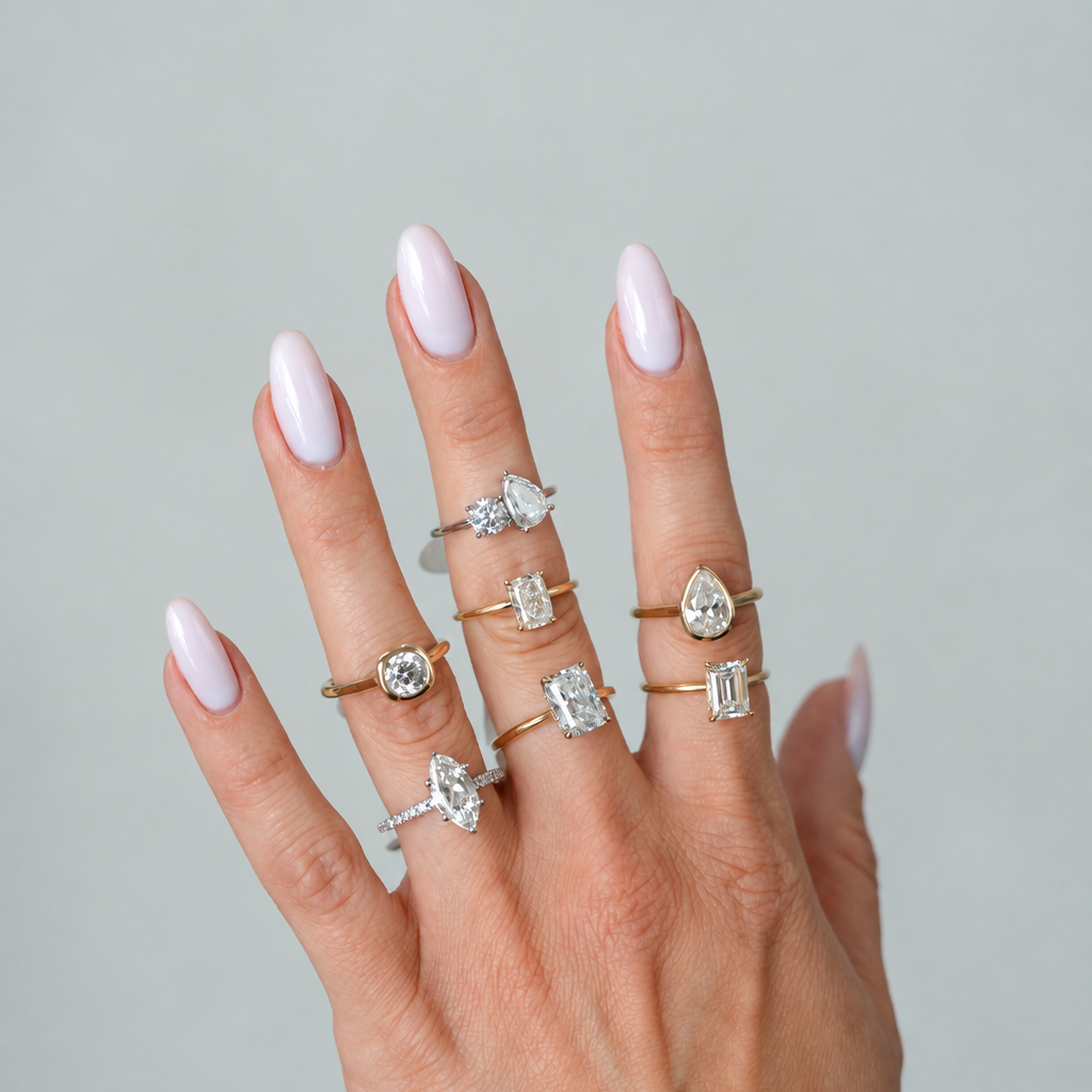 A hand with pale pink manicured nails wearing multiple gold rings with clear gemstones, set against a solid light gray background.